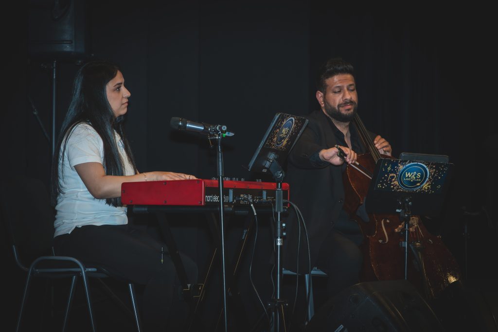 En kvinna spelar keyboard och en man spelar cello på scenen, som har en svart bakgrund och är utrustad med mikrofoner.