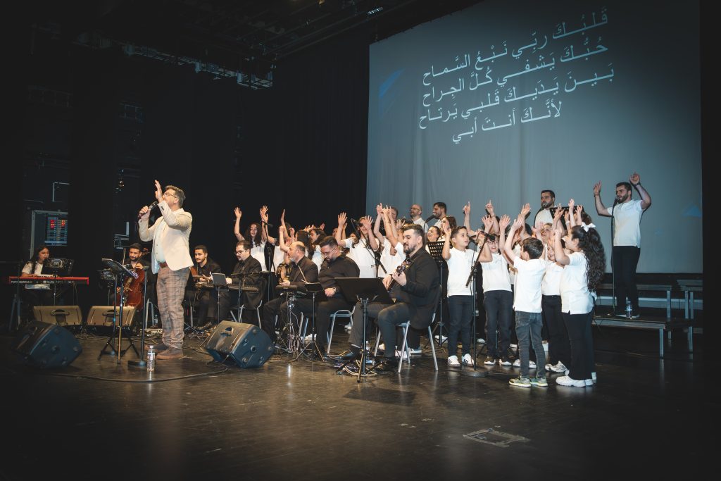 En kör och musiker uppträder på scenen, några med uppsträckta händer; arabiska sångtexter projiceras bakom dem.
