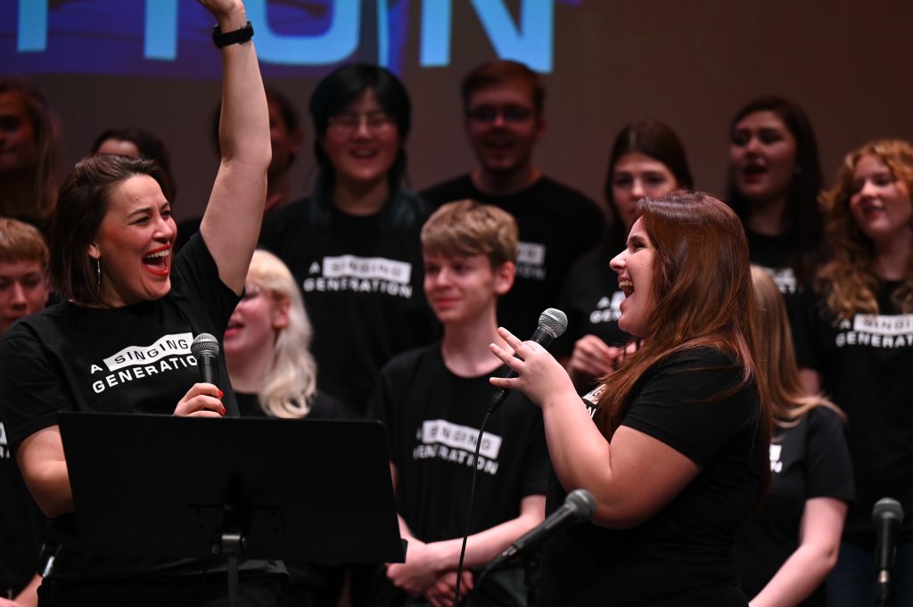 Två kvinnor sjunger glatt på scenen tillsammans med en kör, och alla bär matchande tröjor med texten ”A Singing Generation”.