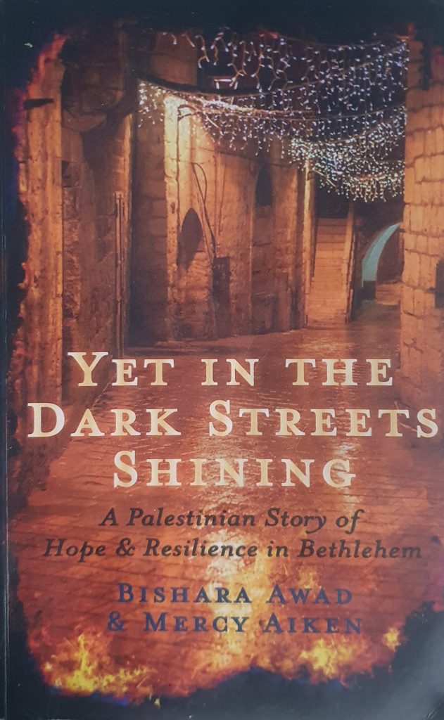 Bokomslag med en stenlagd gränd upplyst av ljusslingor; titeln lyder ”Yet In the Dark Streets Shining.”.