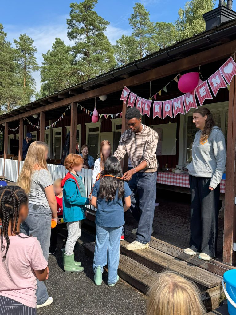 En man räcker över något till en grupp barn utanför en byggnad där det hänger en banderoll med texten ”Välkommen” och ballonger.