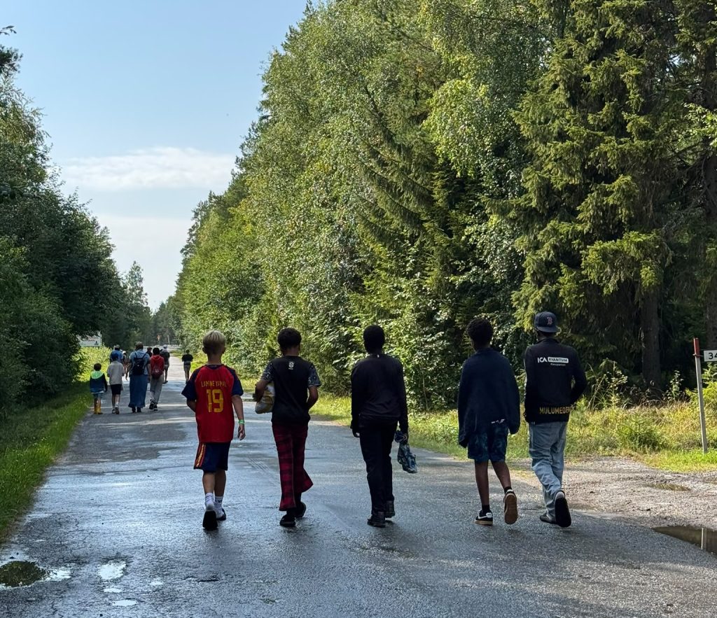 En grupp pojkar går längs en trädkantad väg en solig dag, och i fjärran syns fler människor.