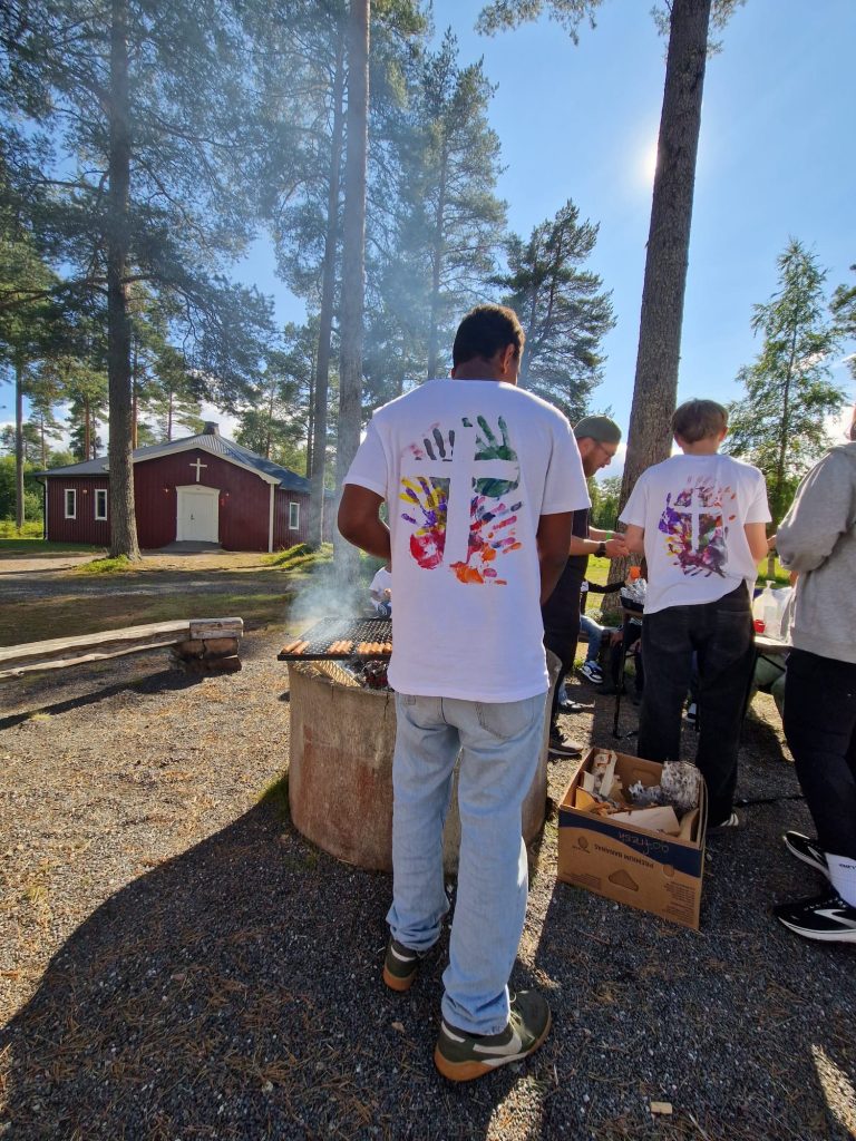 Människor som grillar utomhus nära en röd stuga, klädda i vita skjortor med färgglada handavtrycksmönster.