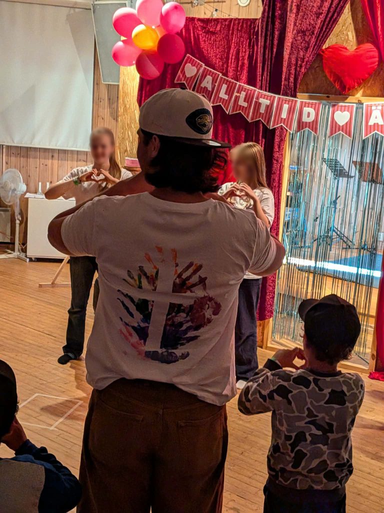 Människor som formar hjärtan med händerna på en träscen, med ballonger och röda hjärtan i bakgrunden.