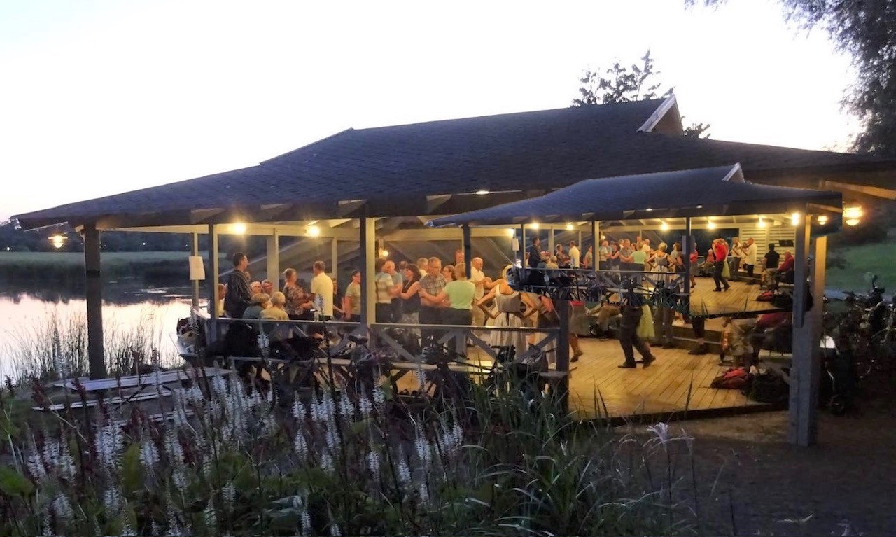 I skymningen samlas människor under en upplyst paviljong vid sjön, umgås och njuter av kvällen tillsammans.