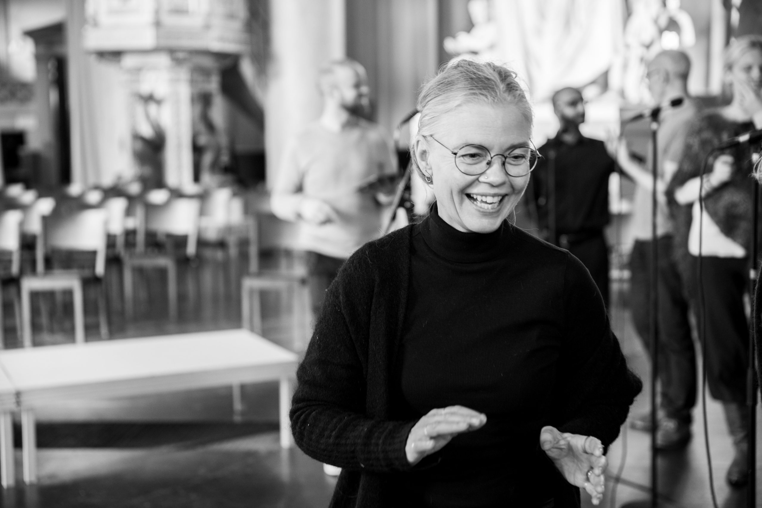 En leende kvinna med glasögon står i fokus; flera personer och stolar är suddiga i bakgrunden.