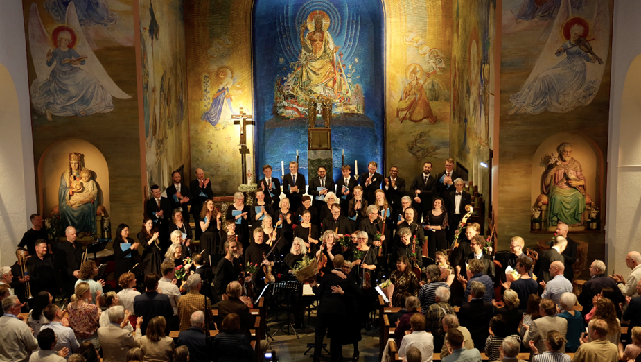 En kör och en orkester uppträder i en kyrka inför en publik, med religiösa väggmålningar i bakgrunden.