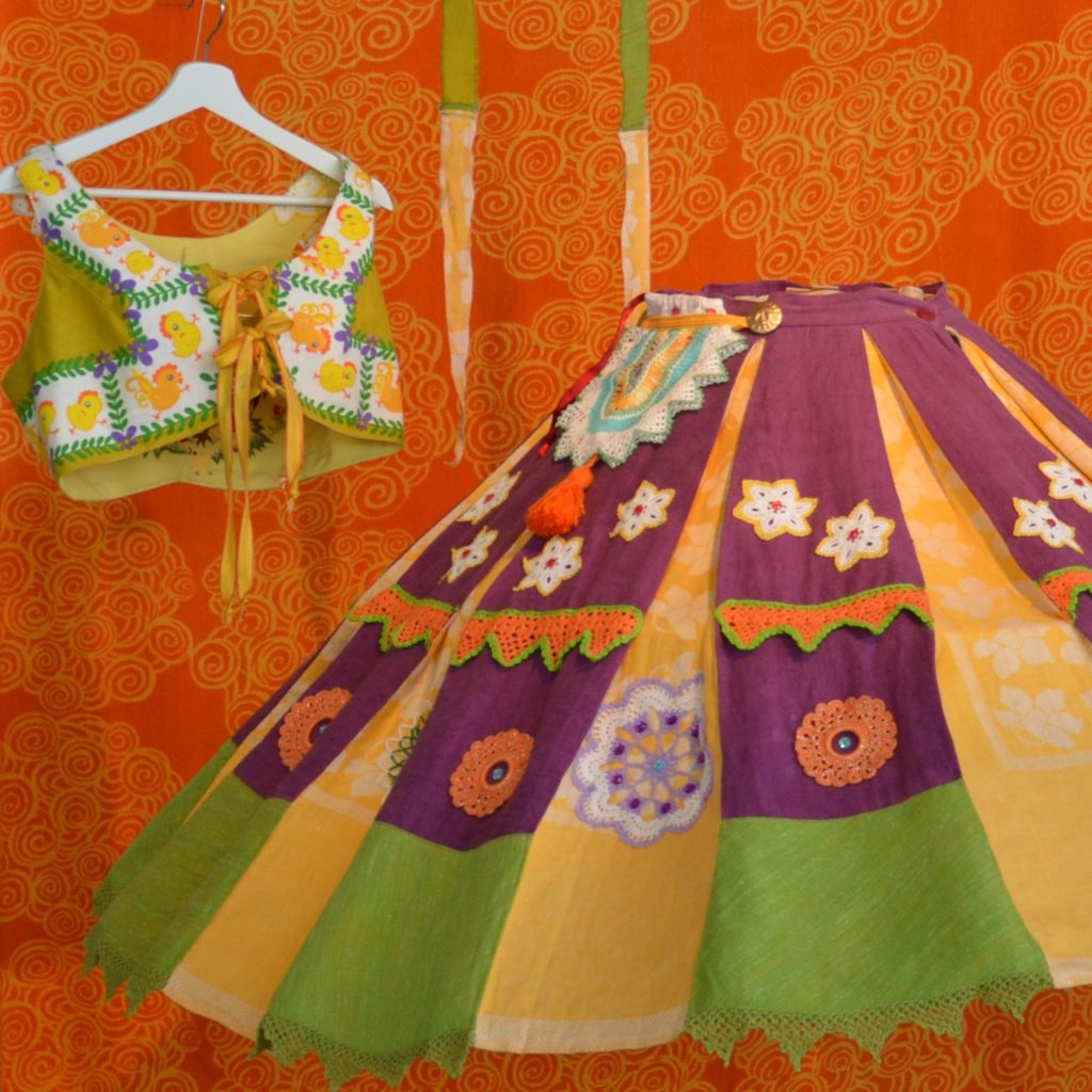 Färgglad traditionell dräkt med broderad överdel och blommig kjol på en orange mönstrad bakgrund.