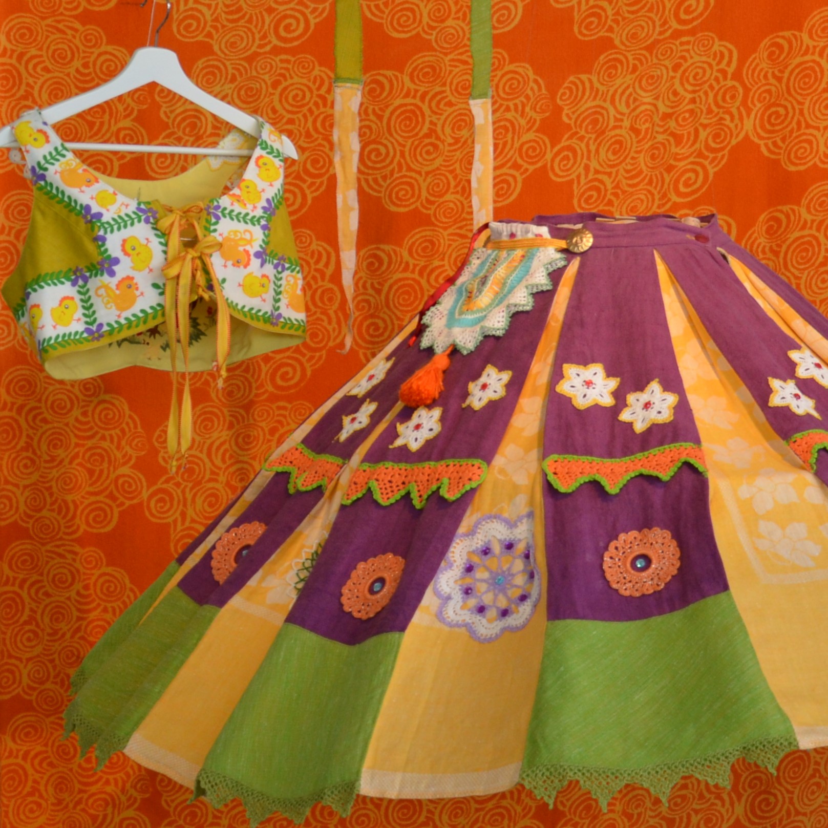 Färgglad traditionell dräkt med broderad överdel och blommig kjol på en orange mönstrad bakgrund.