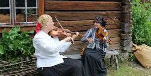 Två kvinnor sitter på en bänk utanför ett trähus, ler och spelar fiol tillsammans.