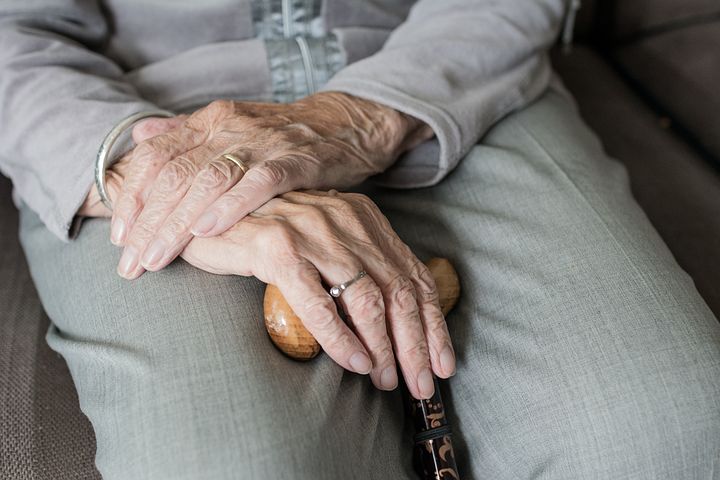 En äldre persons händer vilar på en käpp; personen sitter på en grå soffa och är klädd i grå kläder.