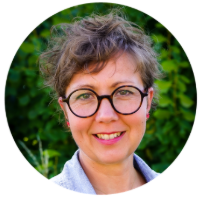 En leende person med kort, lockigt hår och runda glasögon, som står utomhus med grönska i bakgrunden.