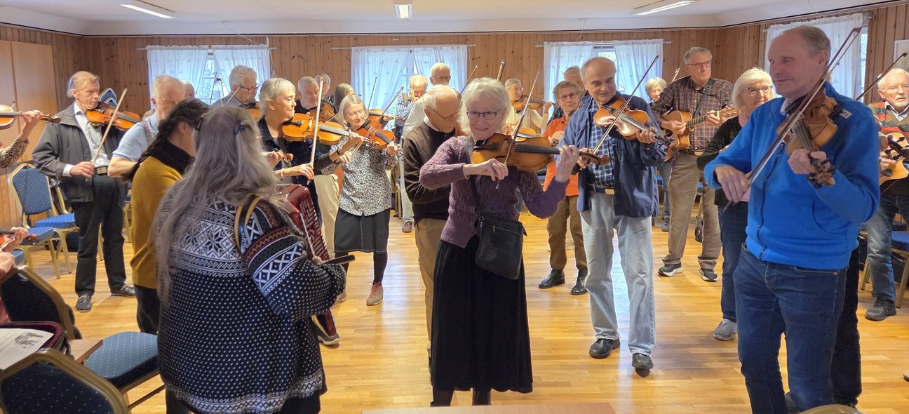 En grupp äldre människor spelar fiol tillsammans i ett ljust rum med träväggar och trästolar.