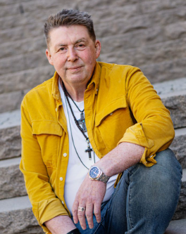 En man i gul skjorta och jeans som sitter på en stentrappa och tittar in i kameran med ett lätt leende.