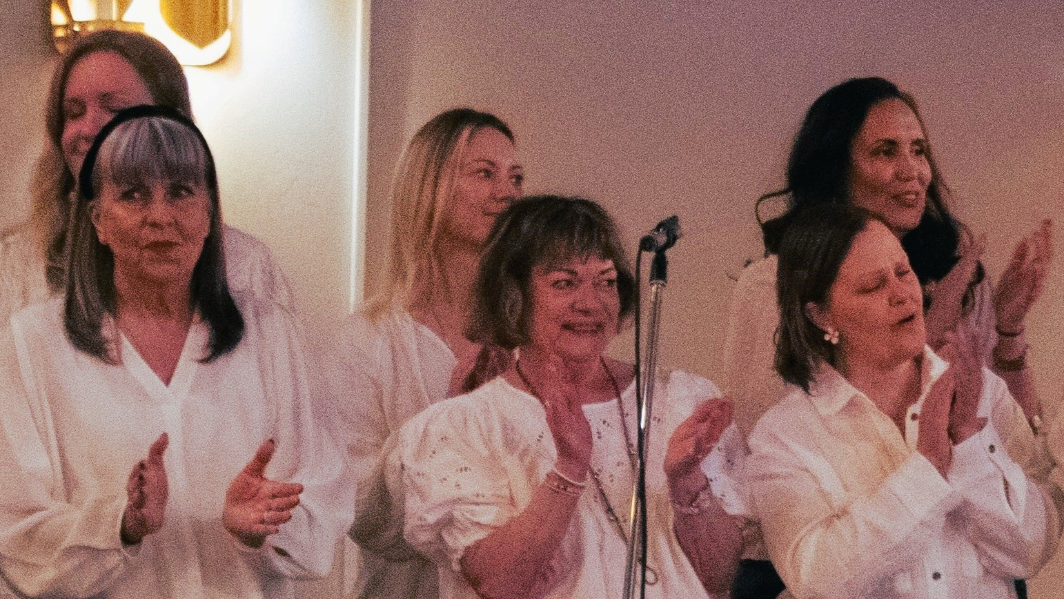 Sex kvinnor i vita skjortor klappar händerna och ler medan de står inomhus, med en mikrofon framför sig.