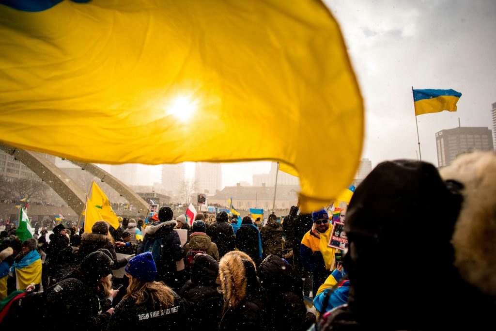 En folkmassa som håller upp ukrainska flaggor vid en demonstration, där solljuset lyser igenom en stor gul och blå flagga.