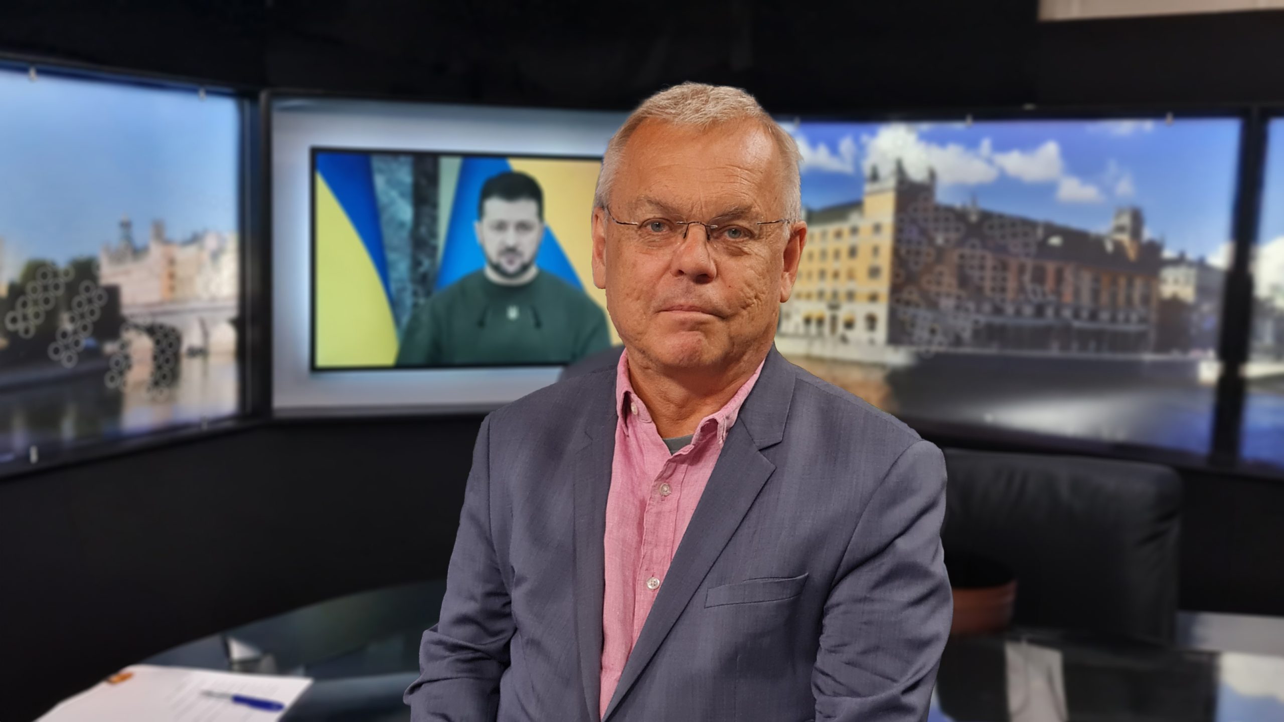 En man med glasögon och kostym står i en TV-studio; på skärmen bakom syns en man mot en stadsbild i bakgrunden.