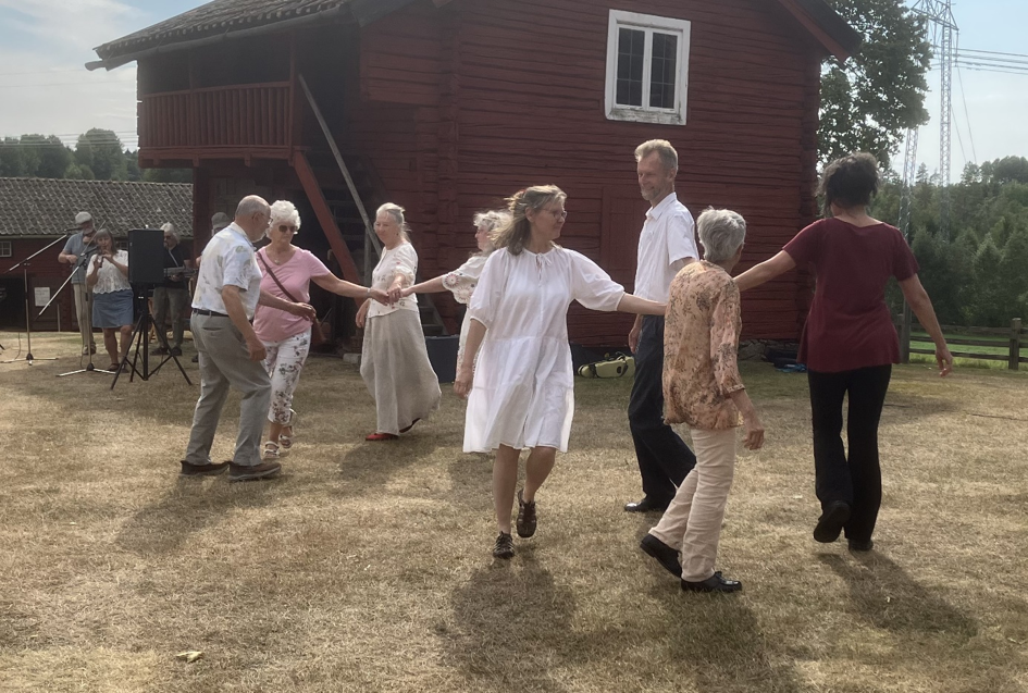 Sju vuxna dansar i en cirkel på torrt gräs nära en röd träbyggnad en solig dag.