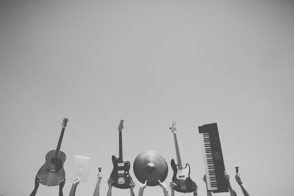 Händerna håller upp en gitarr, en bärbar dator, en cymbal, en elgitarr, en bas, ett keyboard och trumstockar mot himlen.