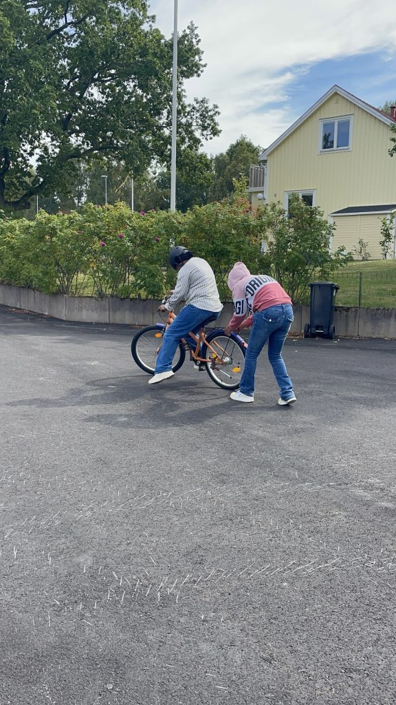 Två personer utomhus; den ena cyklar medan den andra hjälper till genom att hålla i sadeln och skjuta på.