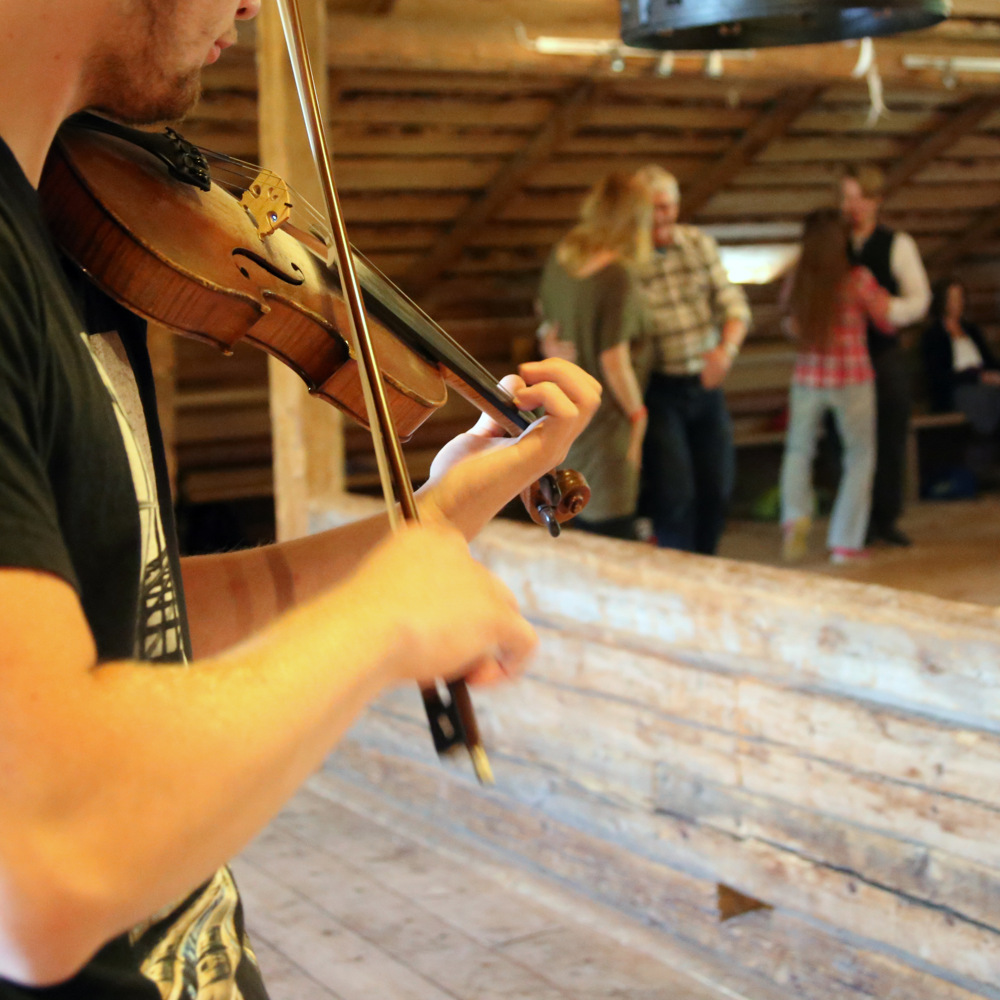 Man spelar fiol och par som dansar folkdans i bakgrunden.