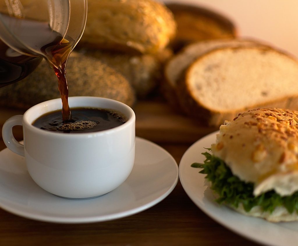 Kaffe hälls upp i en kopp bredvid en smörgås och olika sorters bröd på ett träbord.