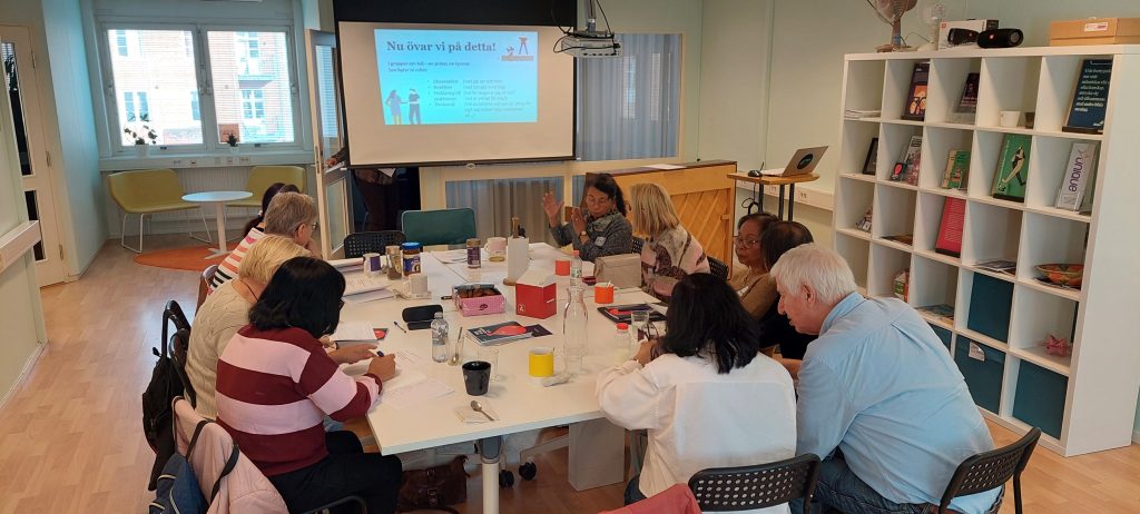 En grupp människor som sitter runt ett bord i ett mötesrum och tittar på en presentation på en skärm.
