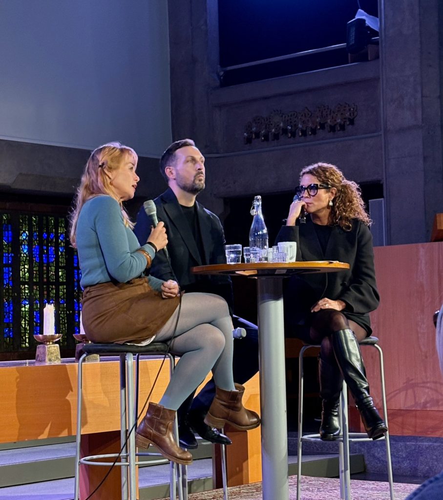 Tre personer sitter på pallar vid ett litet bord under en paneldiskussion.