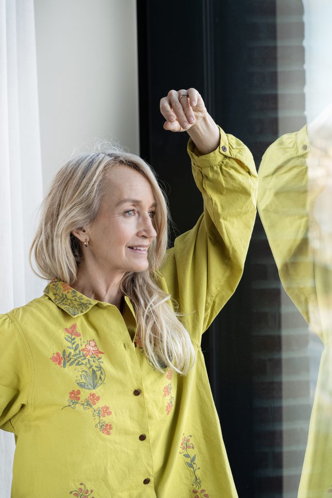 En kvinna i en senapsgul skjorta med blommig brodyr som tittar ut genom ett fönster.