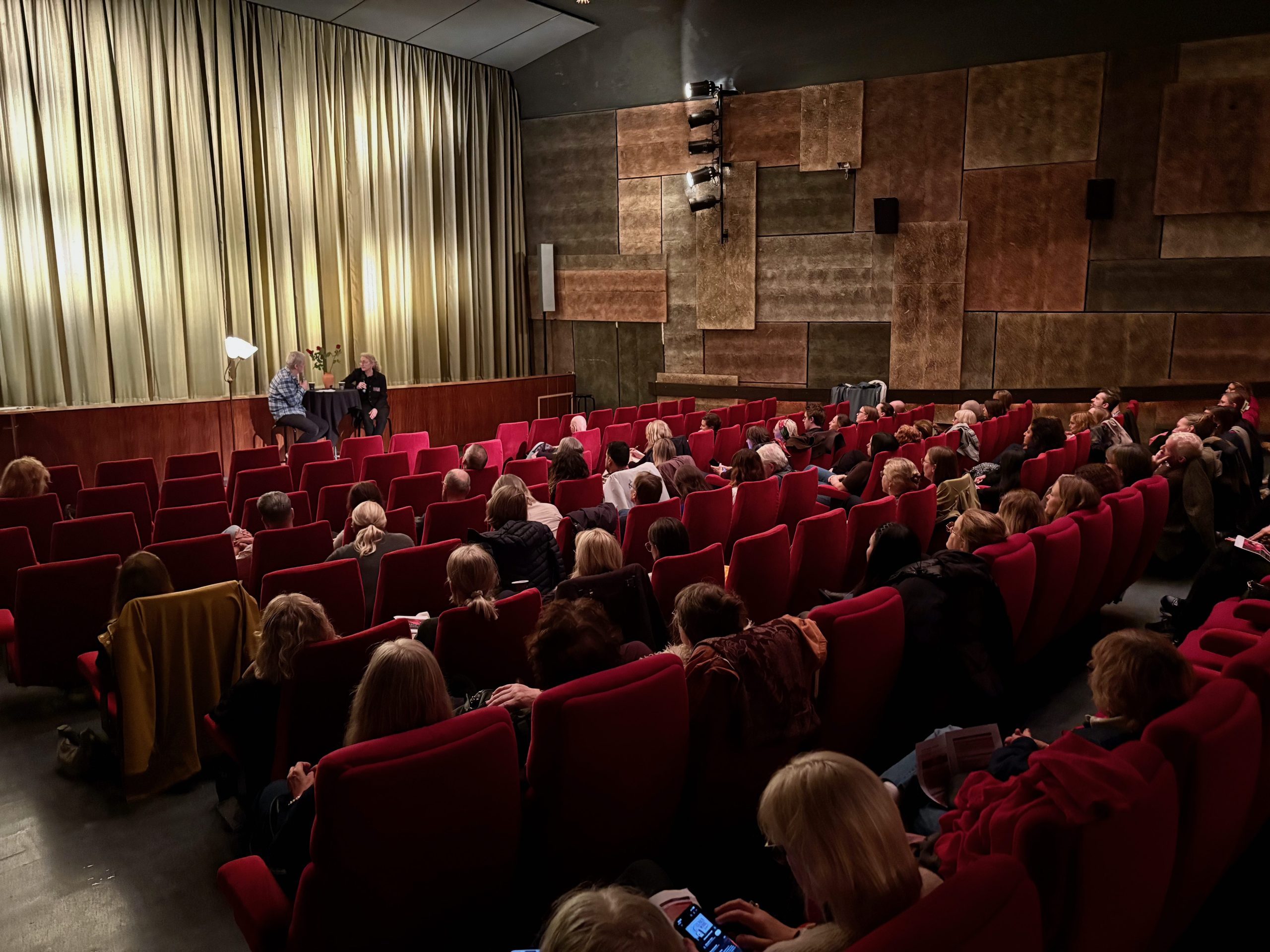 Två talare sitter på scenen framför publiken i en teater med röda stolar.