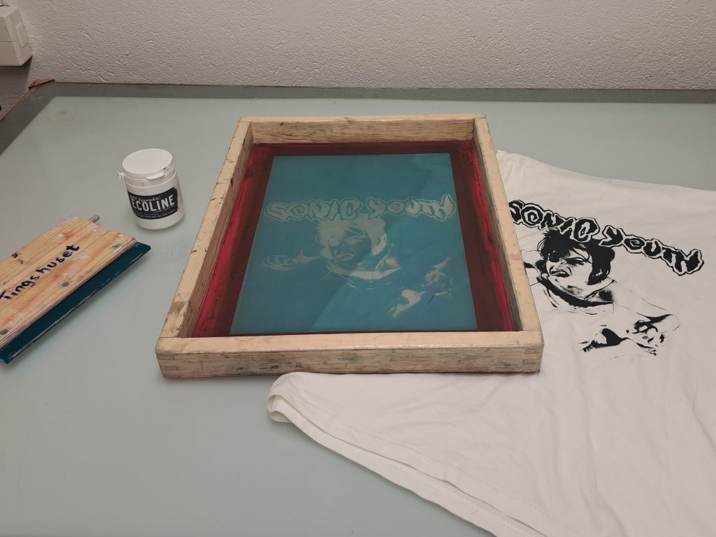 En screentrycksram med ett motiv av Sonic Youth, bredvid en tryckt T-shirt och tryckfärg.