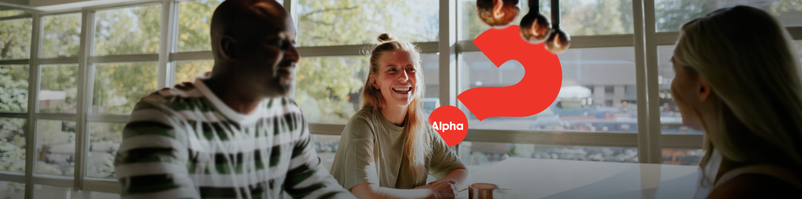 Tre personer som skrattar vid ett bord med en stor röd Alpha-logotyp som överlagring.