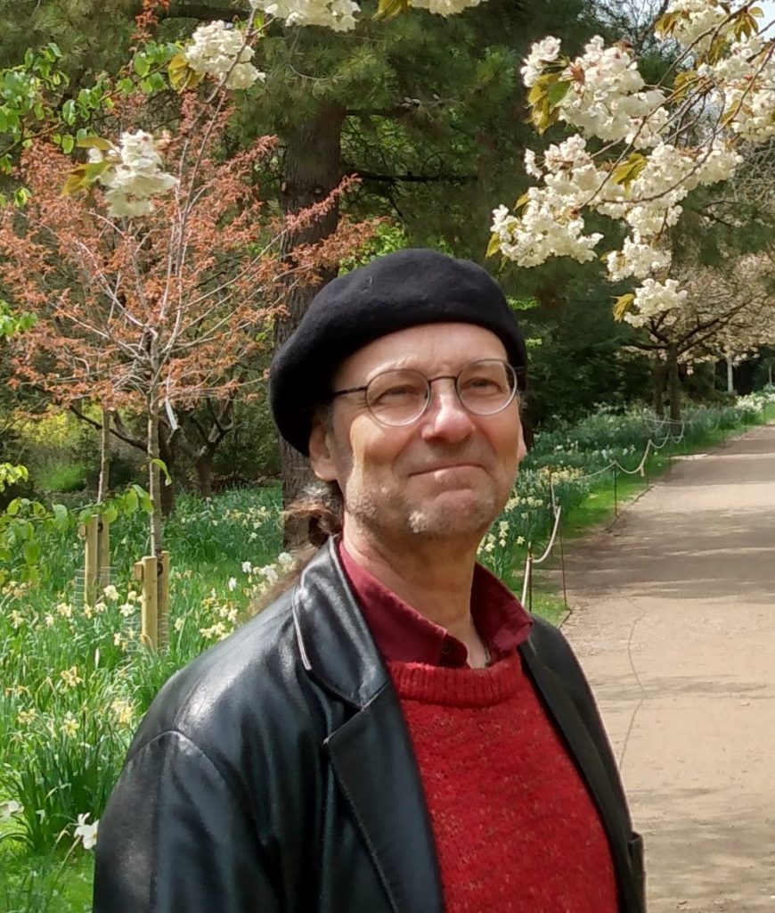 En man med glasögon och basker står på en stig kantad av träd och blommande blommor i en park.