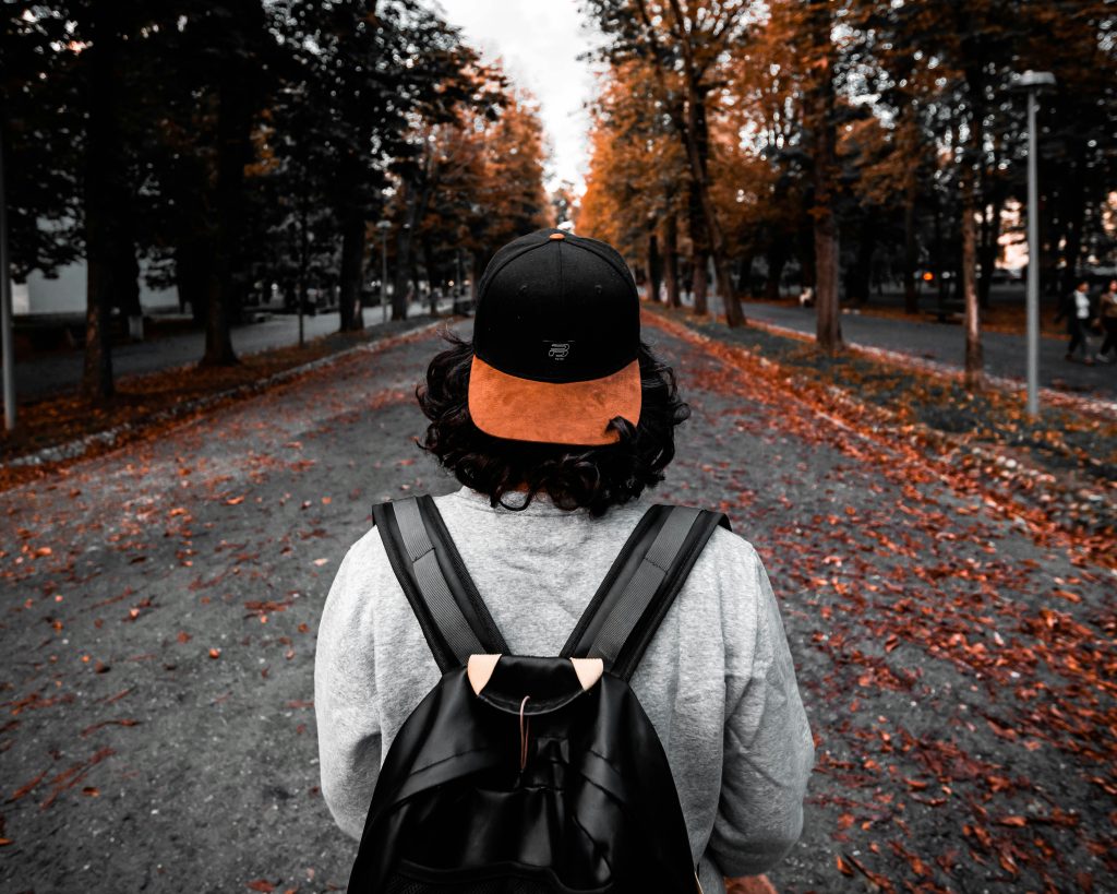 En person med ryggsäck och keps går längs en trädkantad stig som är täckt av orange höstlöv.