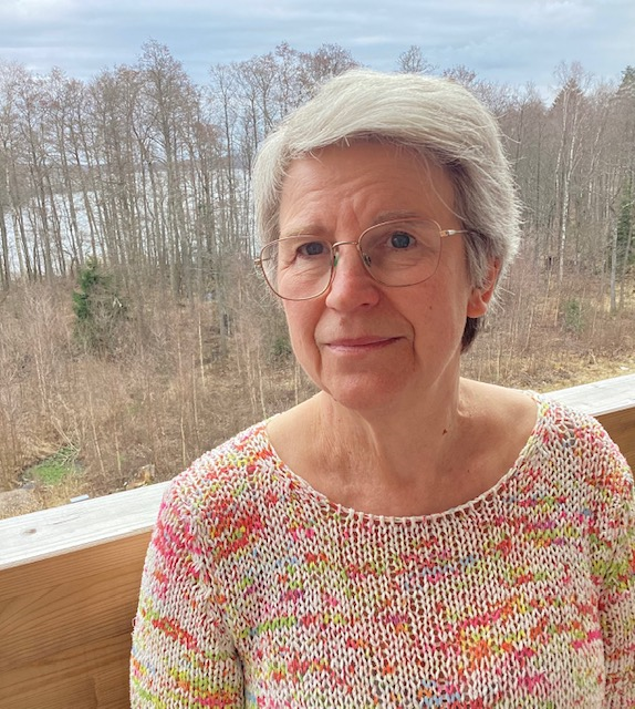 En äldre kvinna med glasögon står på en träbalkong, med träd och en sjö synliga i bakgrunden.